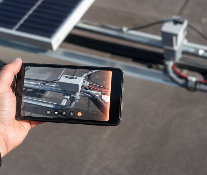 A hand holding a generic smartphone on a rooftop, the screen showing a live video-call view of solar panel wiring and conduit.